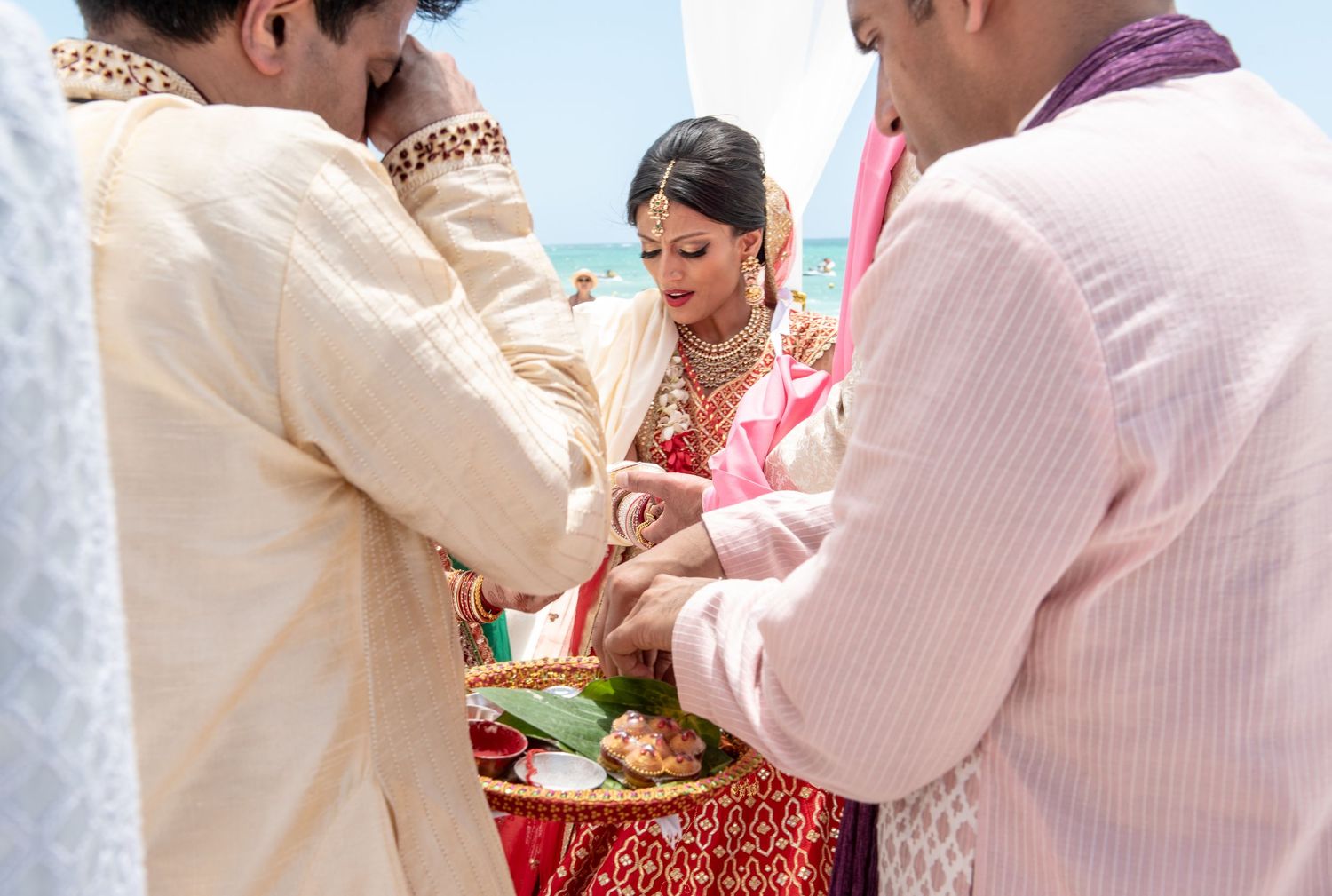 Indian bride in red saree during outdoor beach wedding ceremony with guests participating in traditional rituals.
