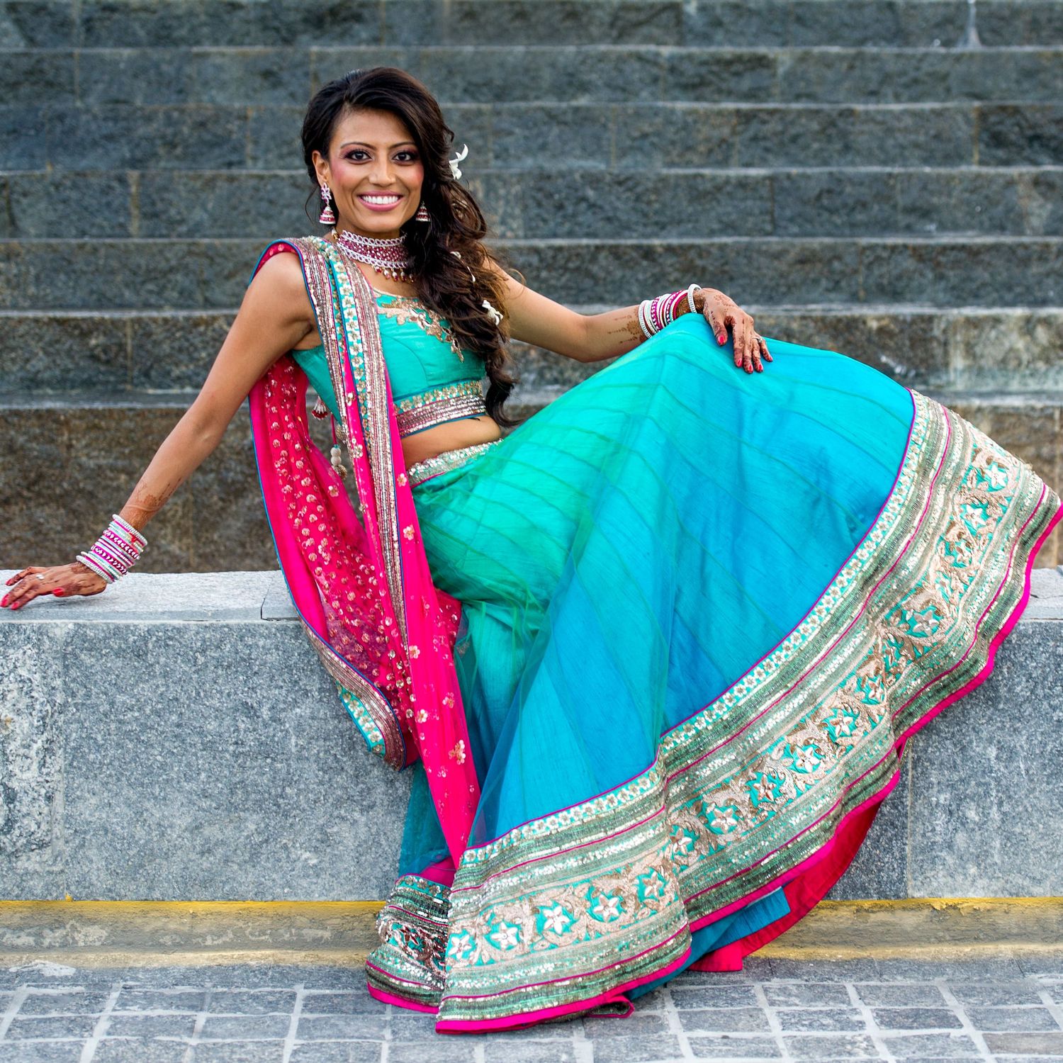 Woman in vibrant turquoise and pink Indian lehenga choli sitting gracefully on stone steps, adorned with traditional jewelry.