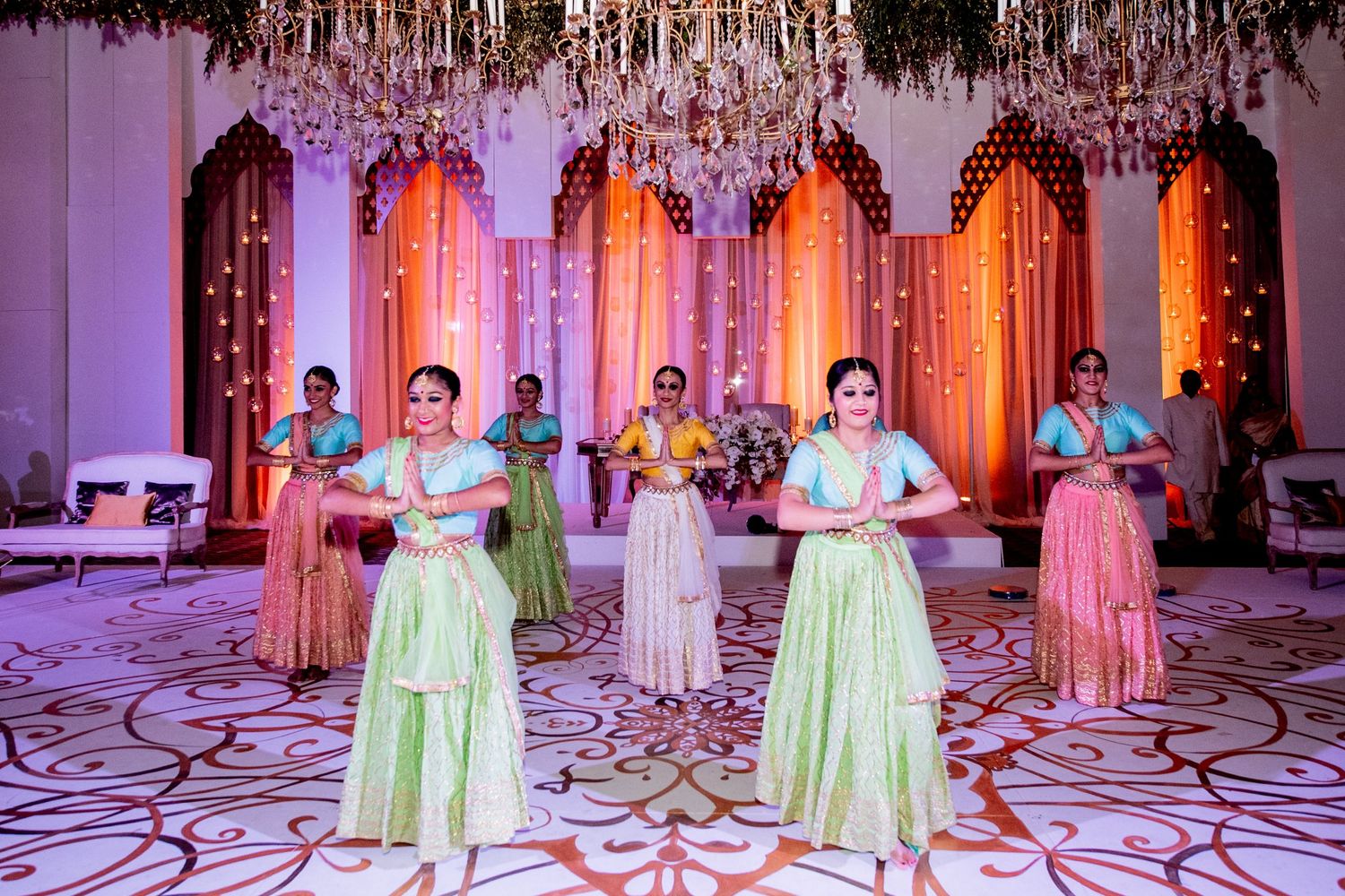 Women in colorful lehengas performing a coordinated dance at an elegantly decorated Indian wedding celebration.