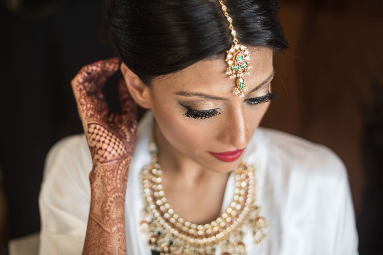 Indian bride in white jacket wearing gold jewelry and maang tikka with intricate henna mehndi on hands.