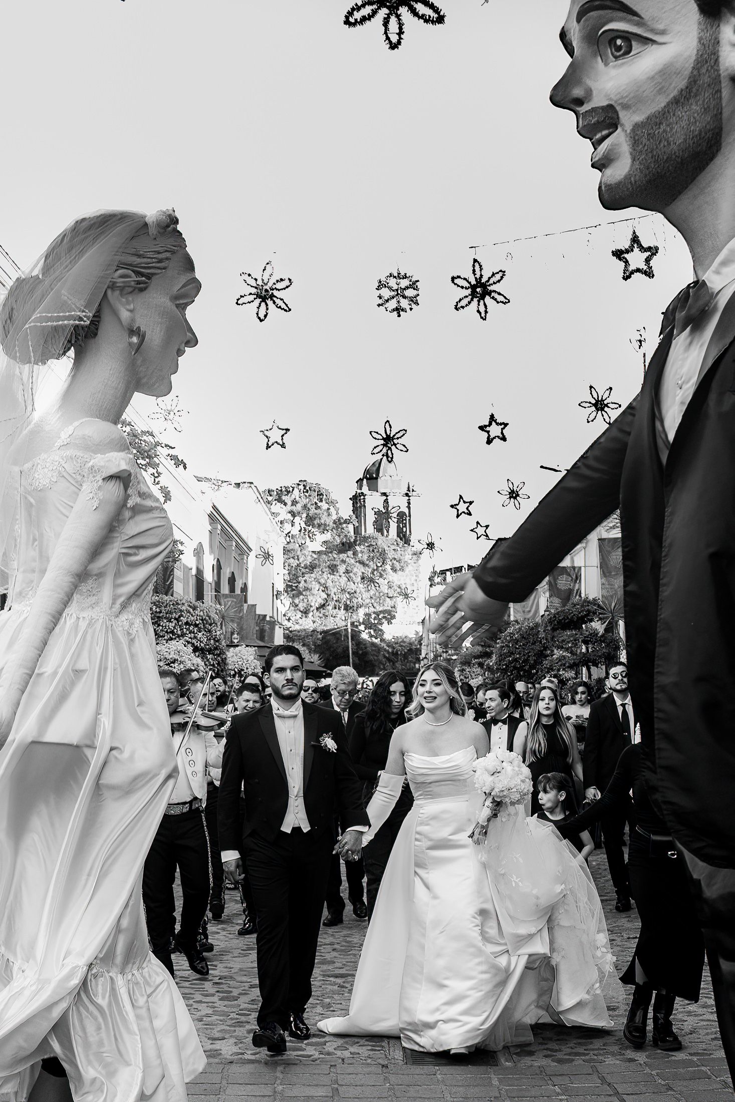 Black and white photo of a bride and groom walking with giant puppet figures during a festive outdoor wedding procession.
