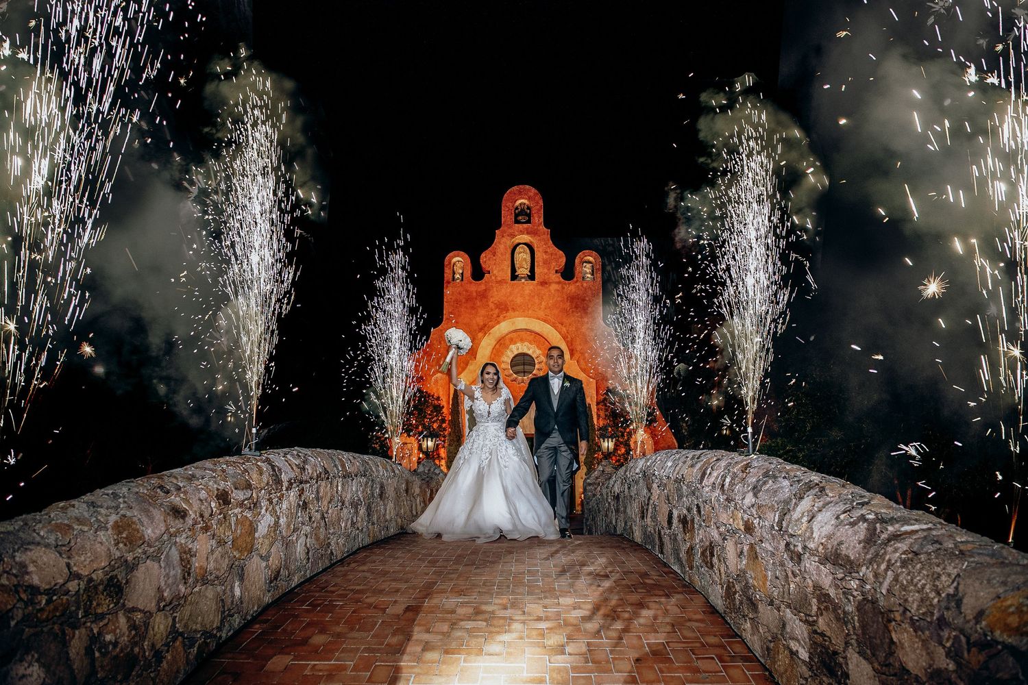 Wedding couple stands on stone bridge with fireworks display in front of illuminated chapel.
