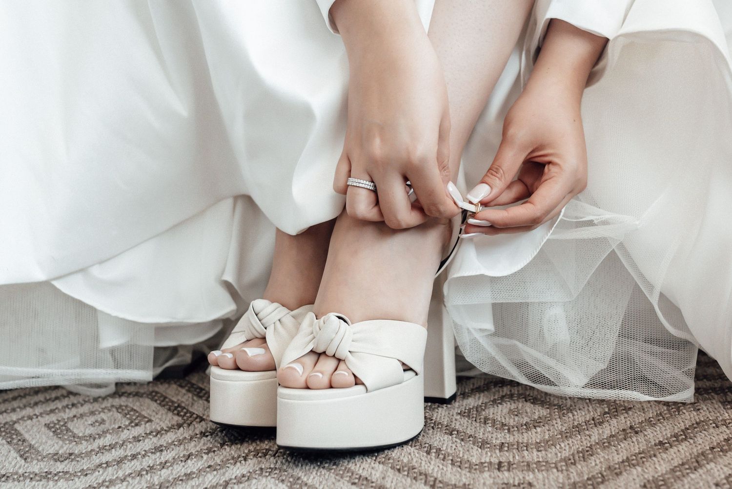 Bride putting on white wedding shoes while wearing elegant white dress.