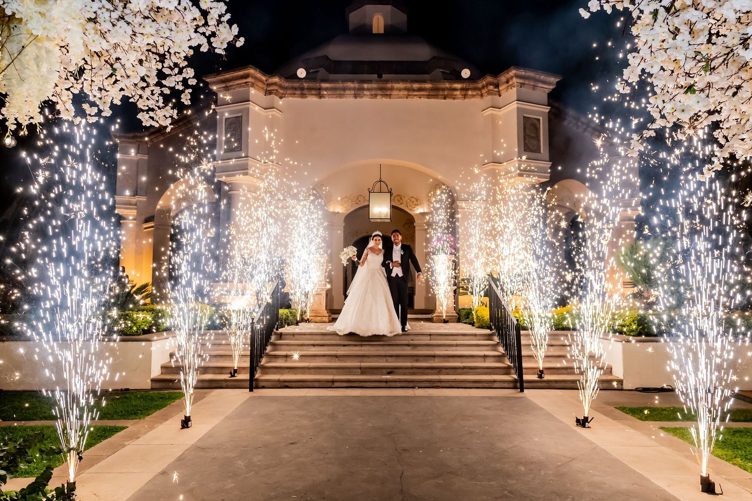 Bride and groom exit grand venue steps flanked by sparkling cold spark fountains and illuminated trees at night.
