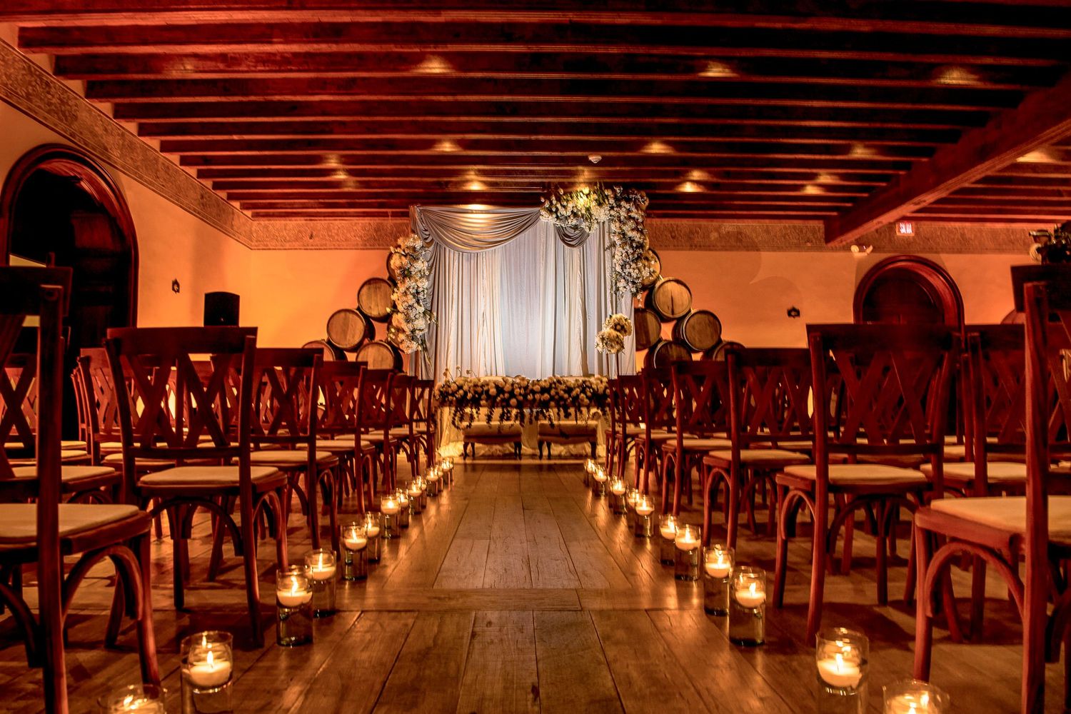Romantic indoor wedding ceremony with candlelit aisle, wooden chairs, floral arch, and warm amber lighting in rustic venue.