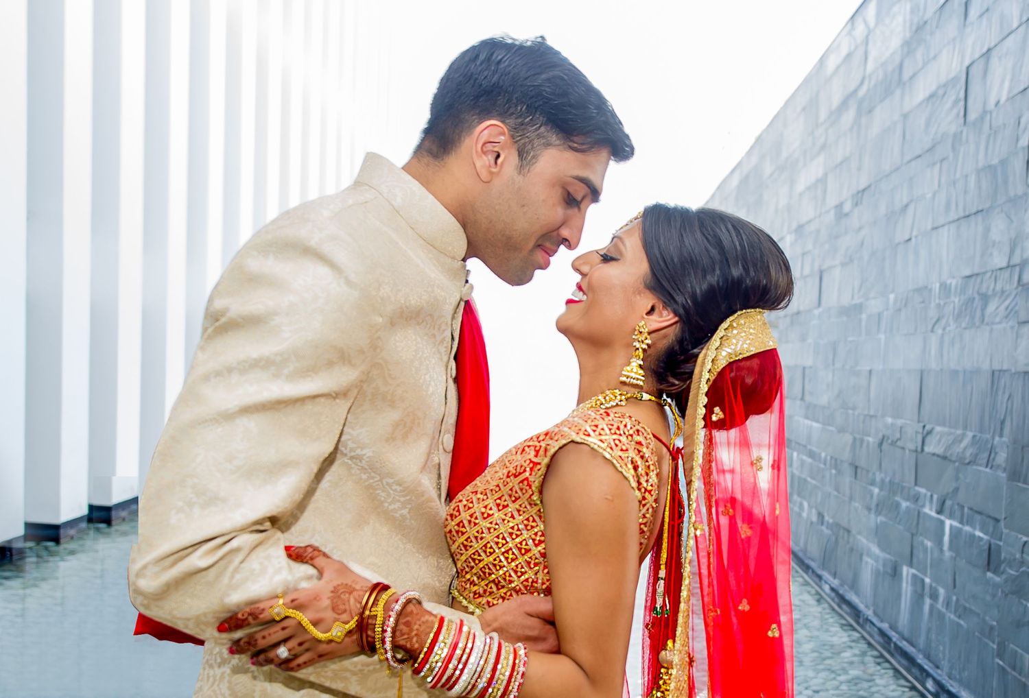 Indian couple in traditional wedding attire share a romantic moment, bride in red saree and groom in cream sherwani.