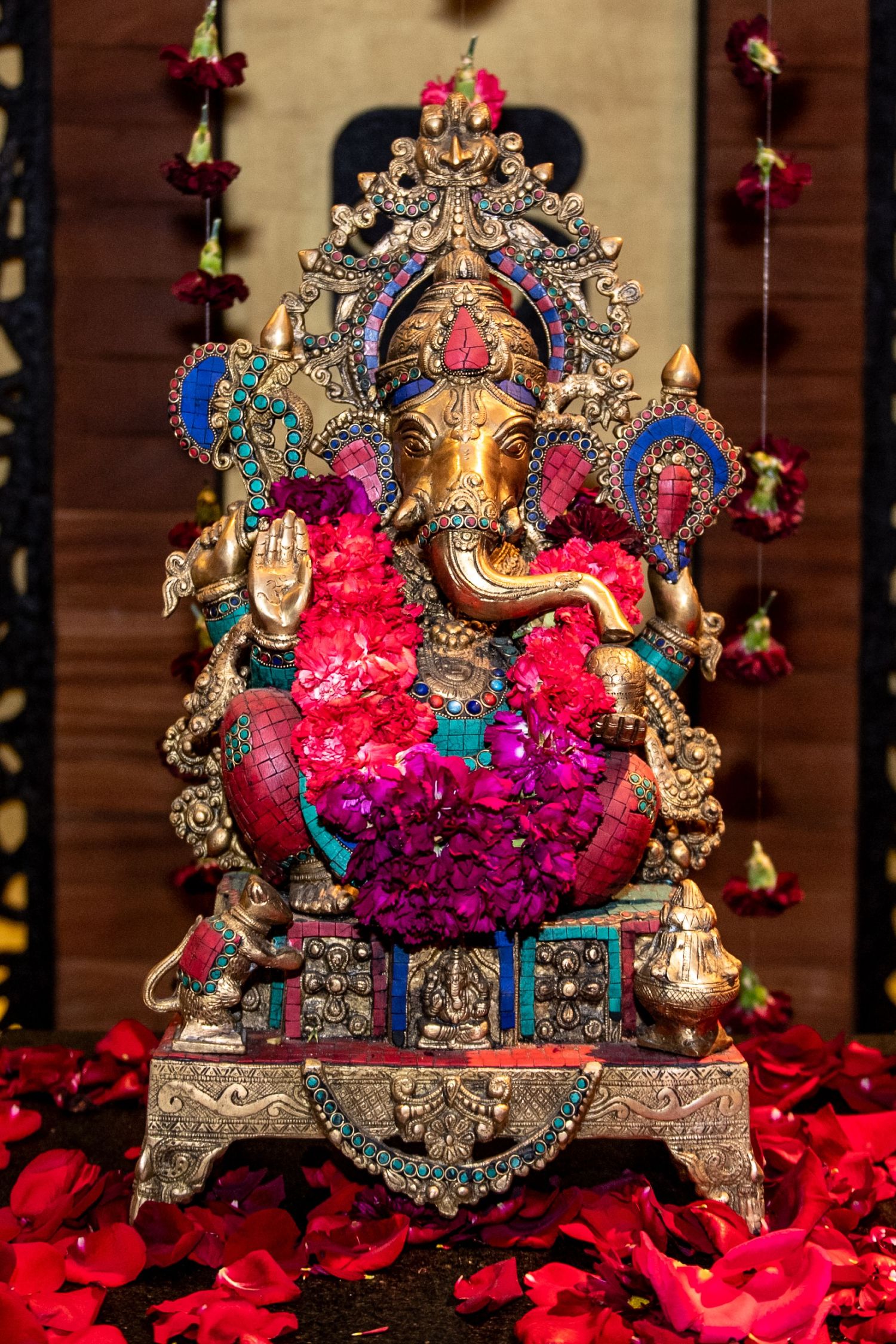 Ornate golden Ganesha idol adorned with vibrant pink flowers and jewels, surrounded by red rose petals on a decorative throne.