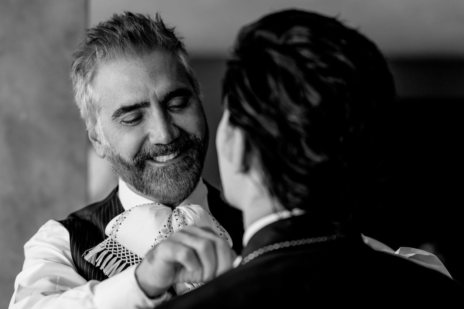 Black and white photo of a smiling bearded man in a vest and tie being assisted by another person.