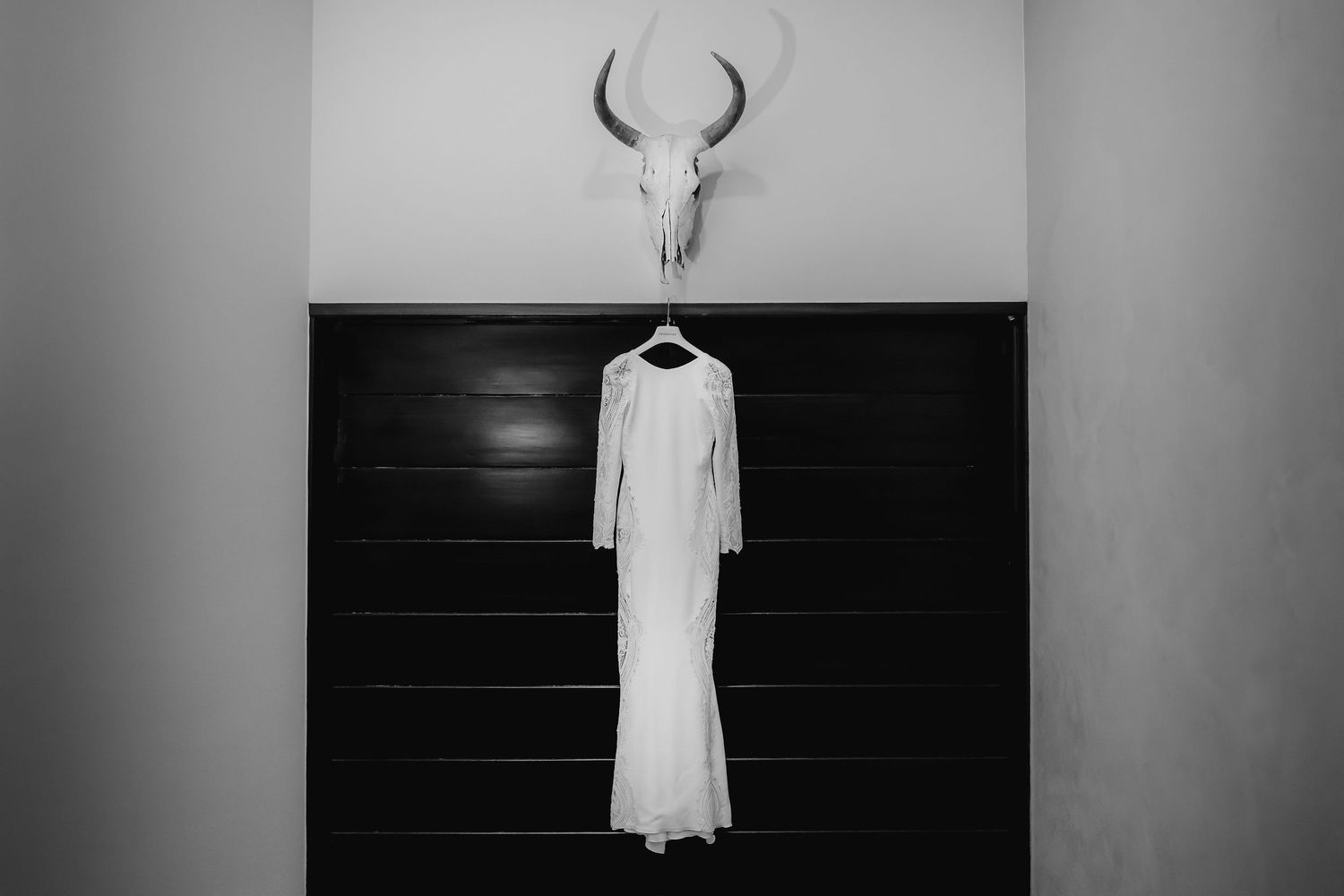 Black and white photo of an elegant white wedding dress hanging on a dark wooden door beneath a deer skull.
