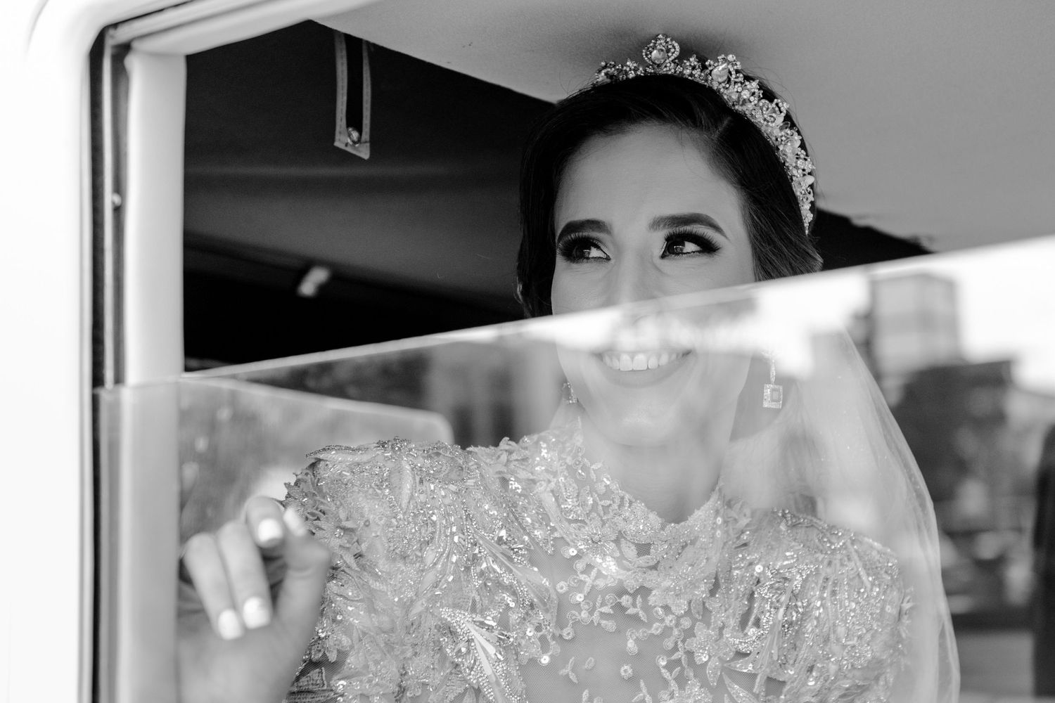 Black and white photo of a smiling bride wearing a tiara and beaded gown, looking up while seated in a vehicle.
