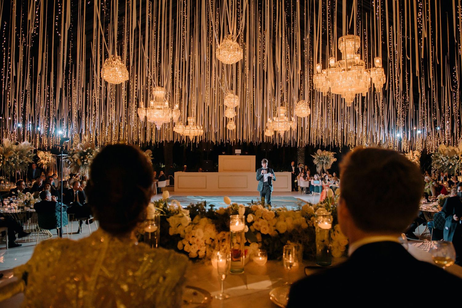 Elegant wedding reception with chandelier ceiling, hanging gold strands, floral centerpieces, and couple on dance floor.