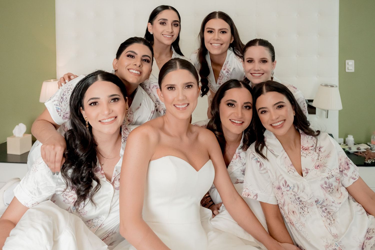 Bride in white strapless gown poses with seven bridesmaids in floral robes, smiling together in a bright room.