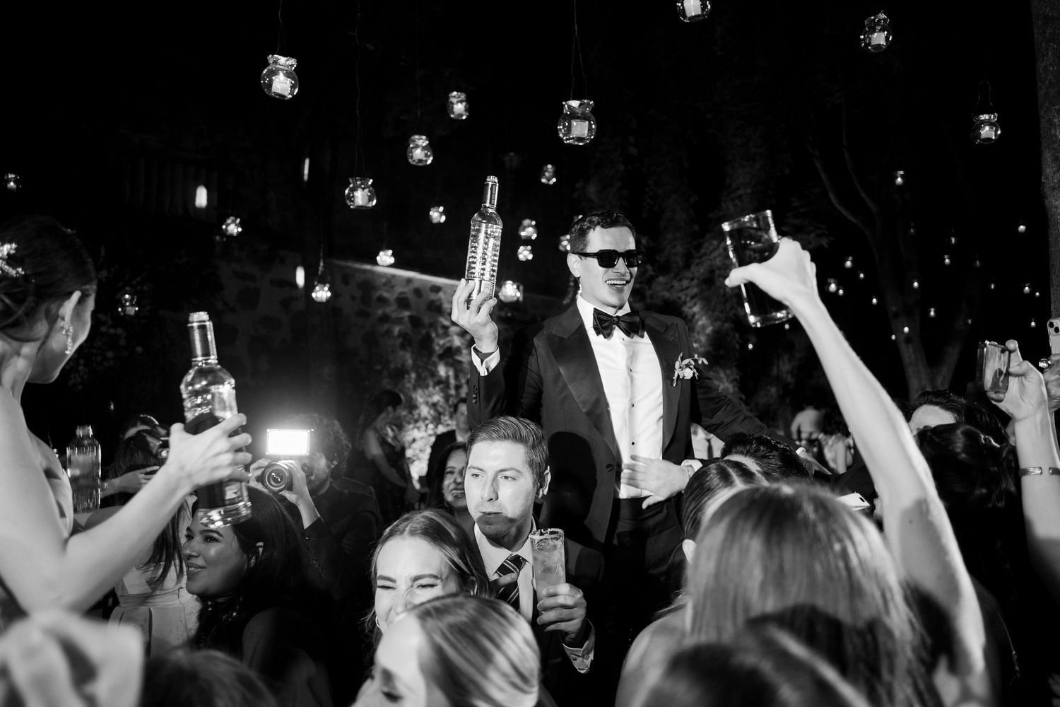 Black and white photo of groom in tuxedo crowd surfing at wedding reception party with guests raising drinks.