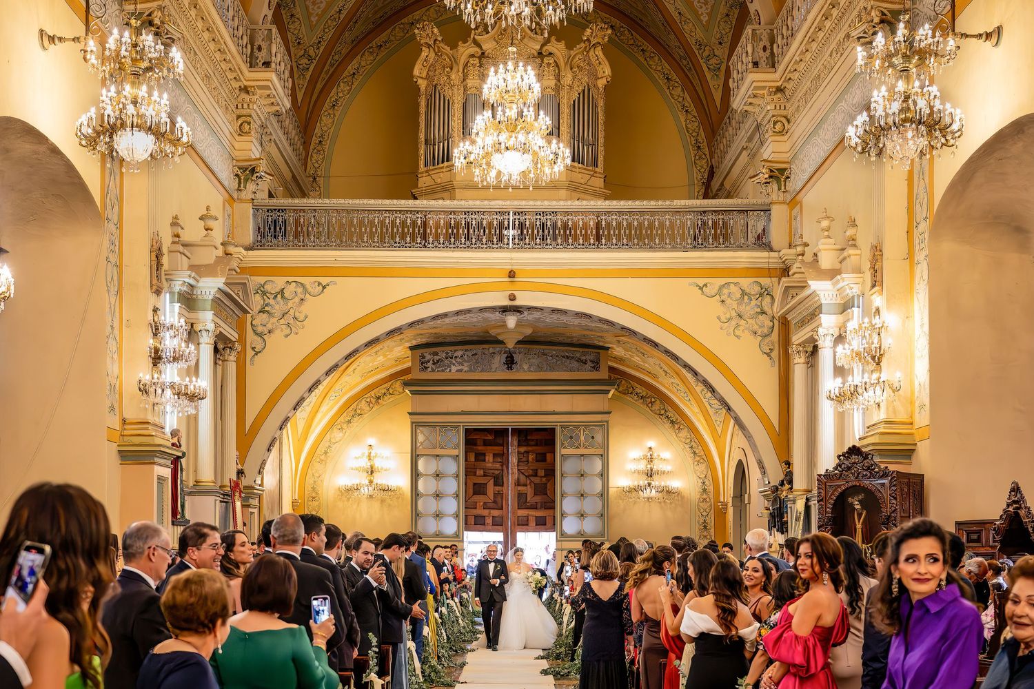 Elegant wedding ceremony in ornate golden hall with chandeliers, guests lining aisle as bride walks toward grand wooden doors.