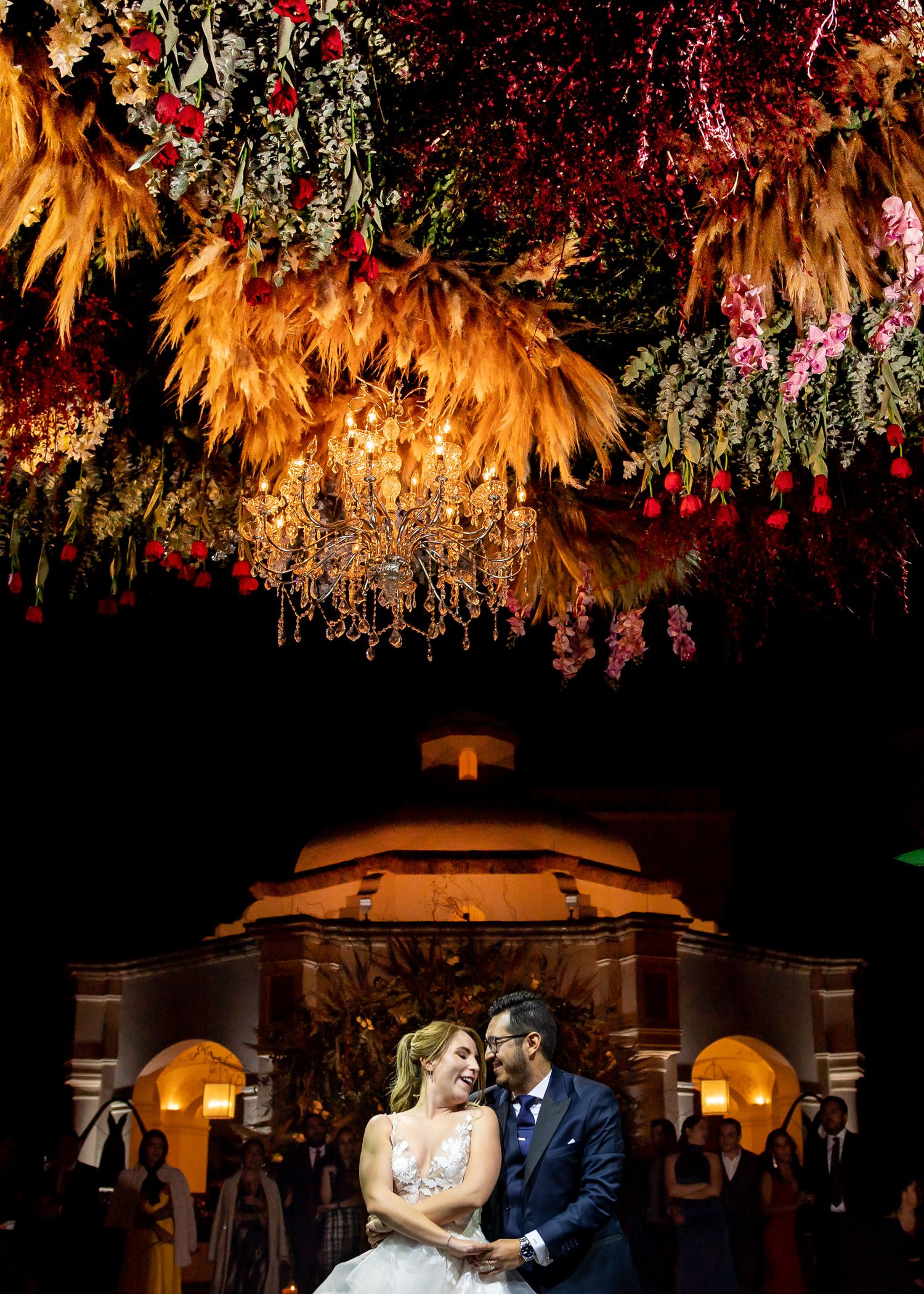 Bride and groom laughing under stunning floral ceiling installation with golden palms and red blooms at elegant night wedding.