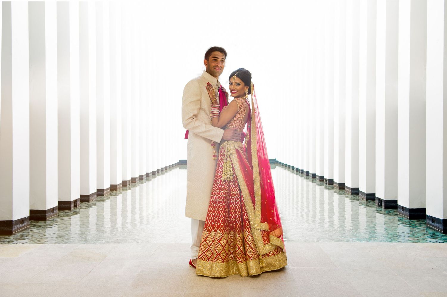 Indian couple in traditional wedding attire posing elegantly between white columns near a reflective pool.