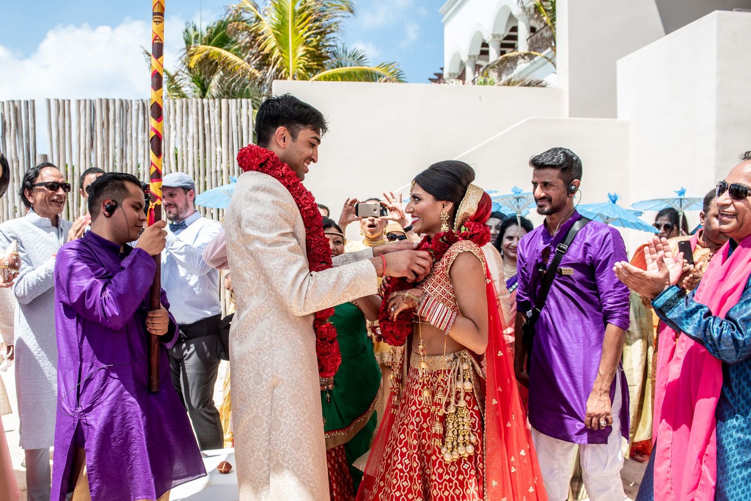 Vibrant Indian wedding ceremony with bride in red lehenga and groom in white sherwani exchanging garlands outdoors.