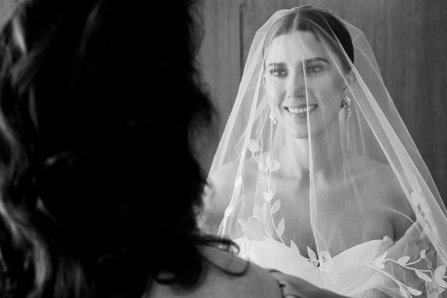 Black and white photo of a smiling bride wearing a veil, seen in a mirror reflection on her wedding day.