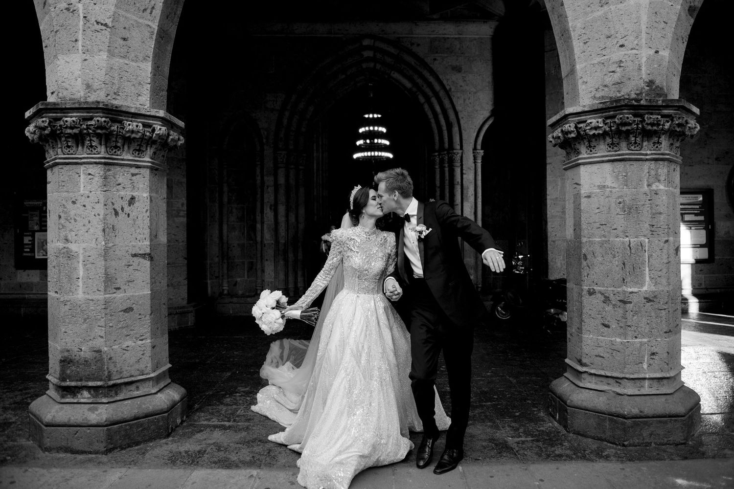 Black and white photo of a bride and groom sharing a kiss under stone arches at a romantic wedding venue.