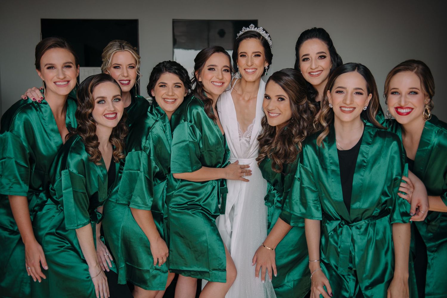 Bride in white robe poses with bridesmaids wearing matching emerald green satin robes before wedding ceremony.