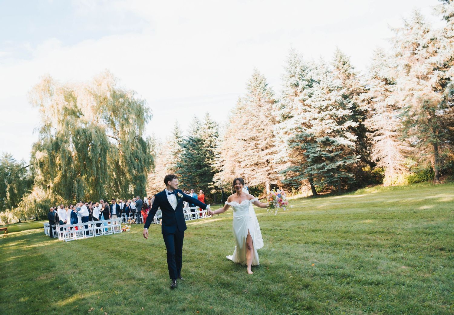 Wedding ceremony setup in a scenic outdoor garden with white chairs arranged in rows beneath tall trees.