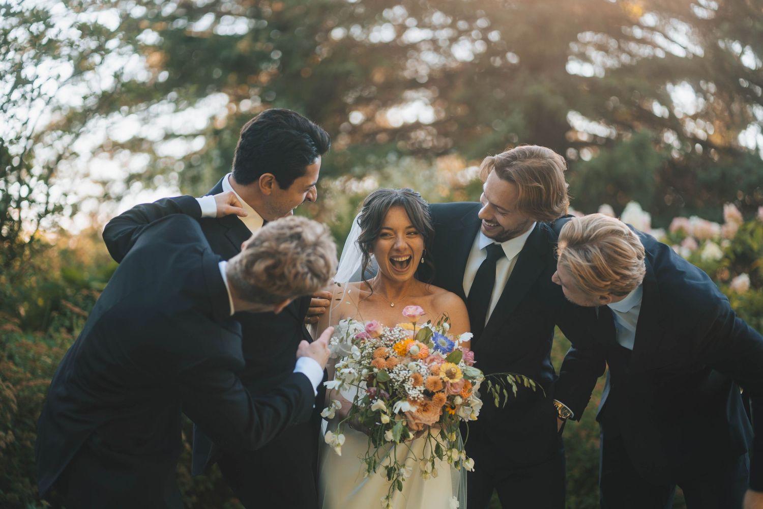 A romantic wedding couple embraces outdoors during golden hour while guests celebrate in the background.