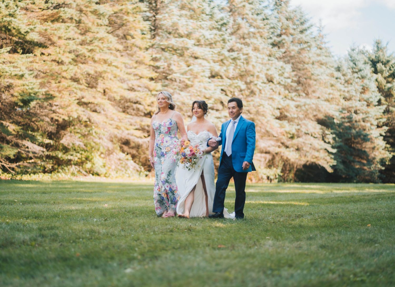 A wedding party walks across a sunlit lawn with evergreen trees and golden foliage in the background.