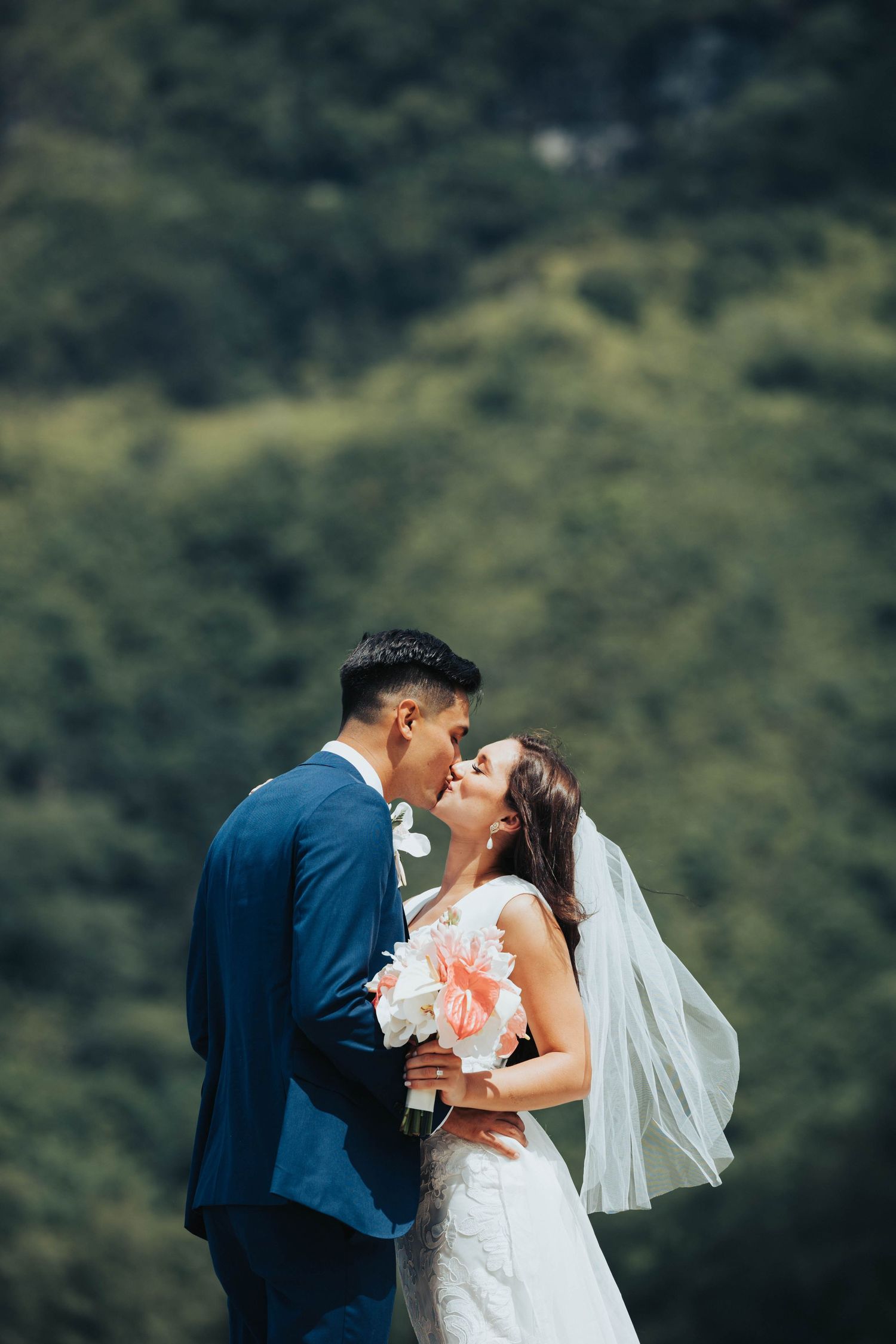 A romantic wedding couple shares a kiss outdoors with lush green mountains in the background.