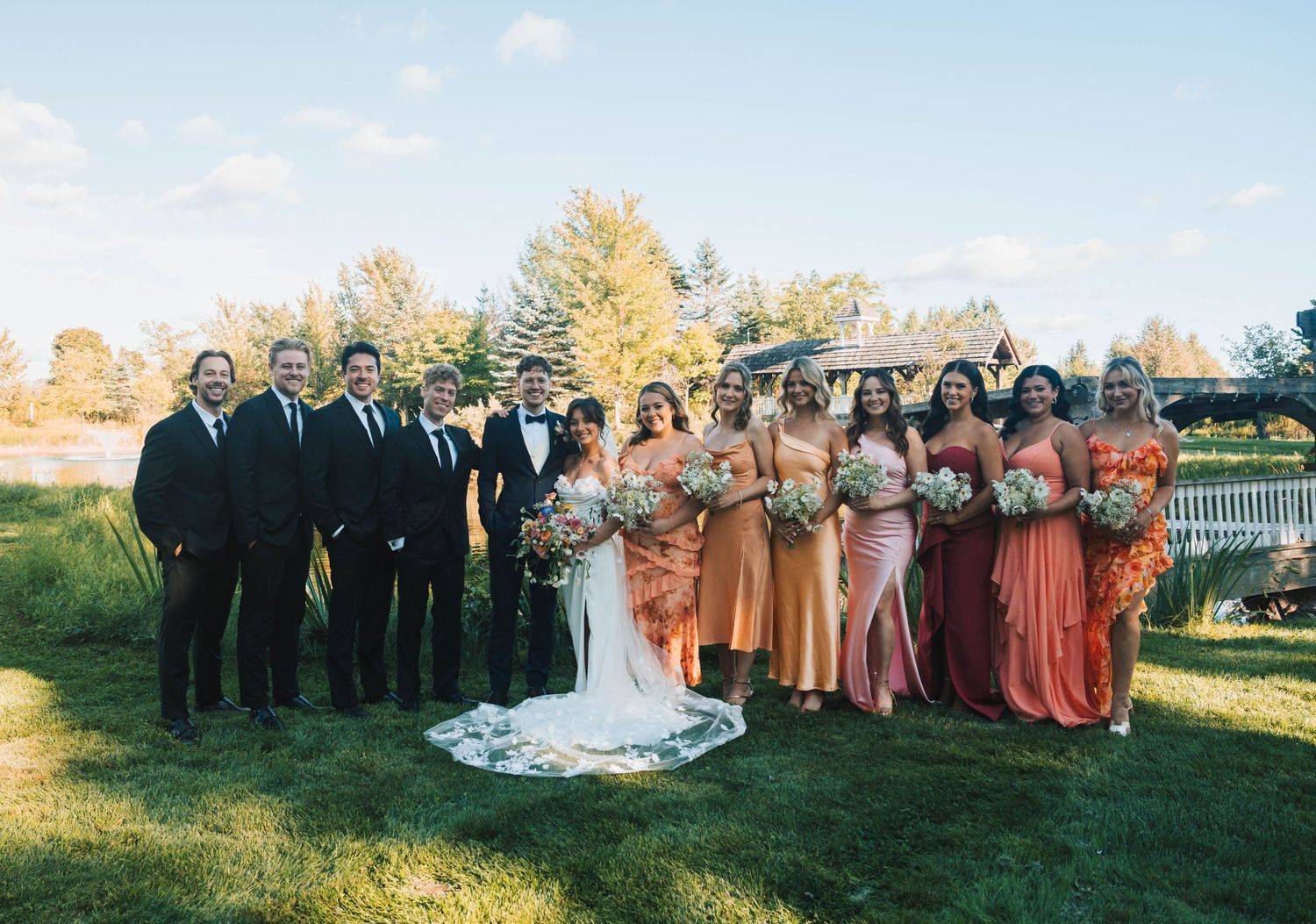 Wedding party poses outdoors in autumn with bridesmaids in rust and coral dresses and groomsmen in black suits.