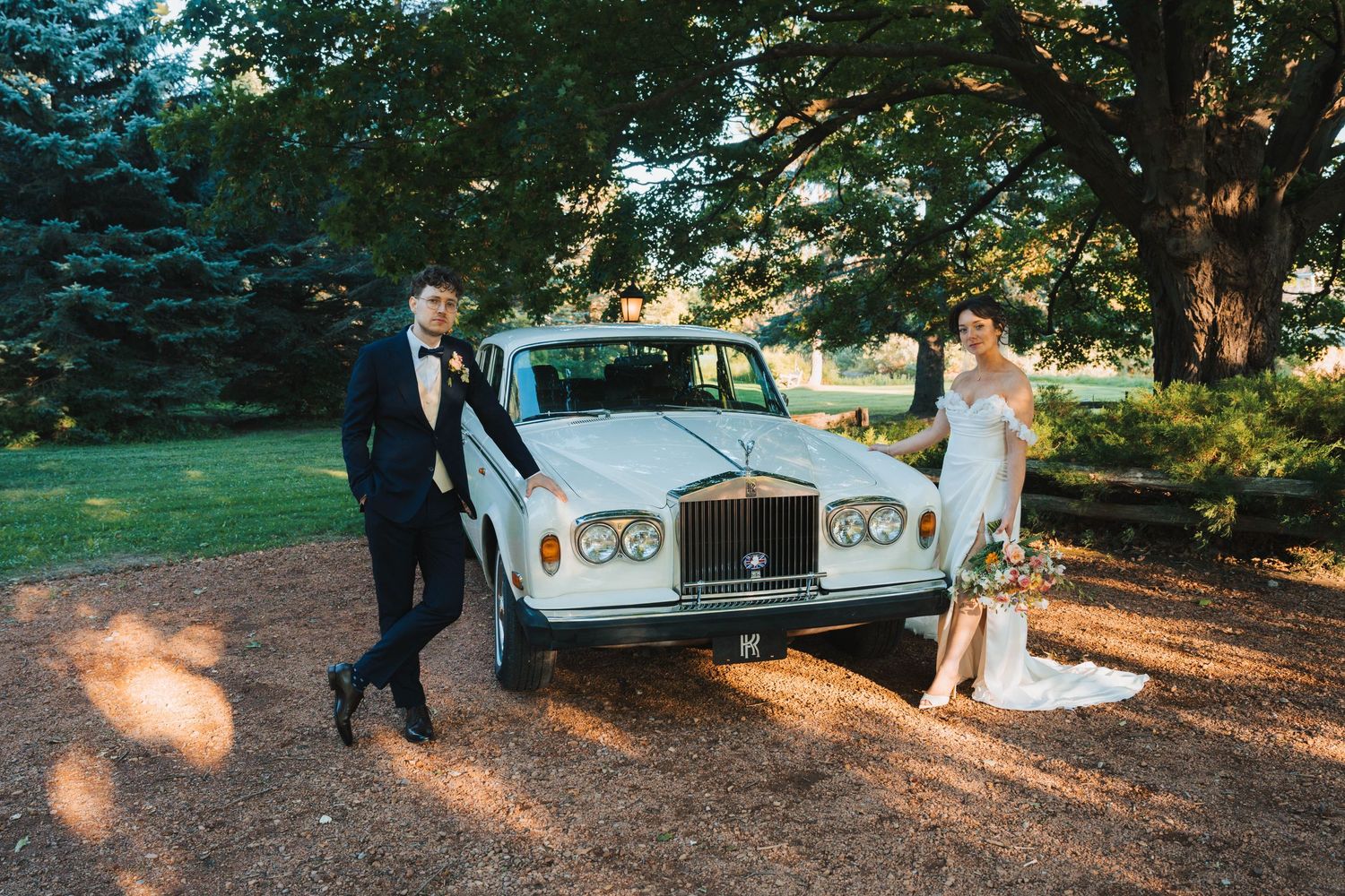A couple walks together through a sunlit forest path with a vintage white Rolls Royce in the background.