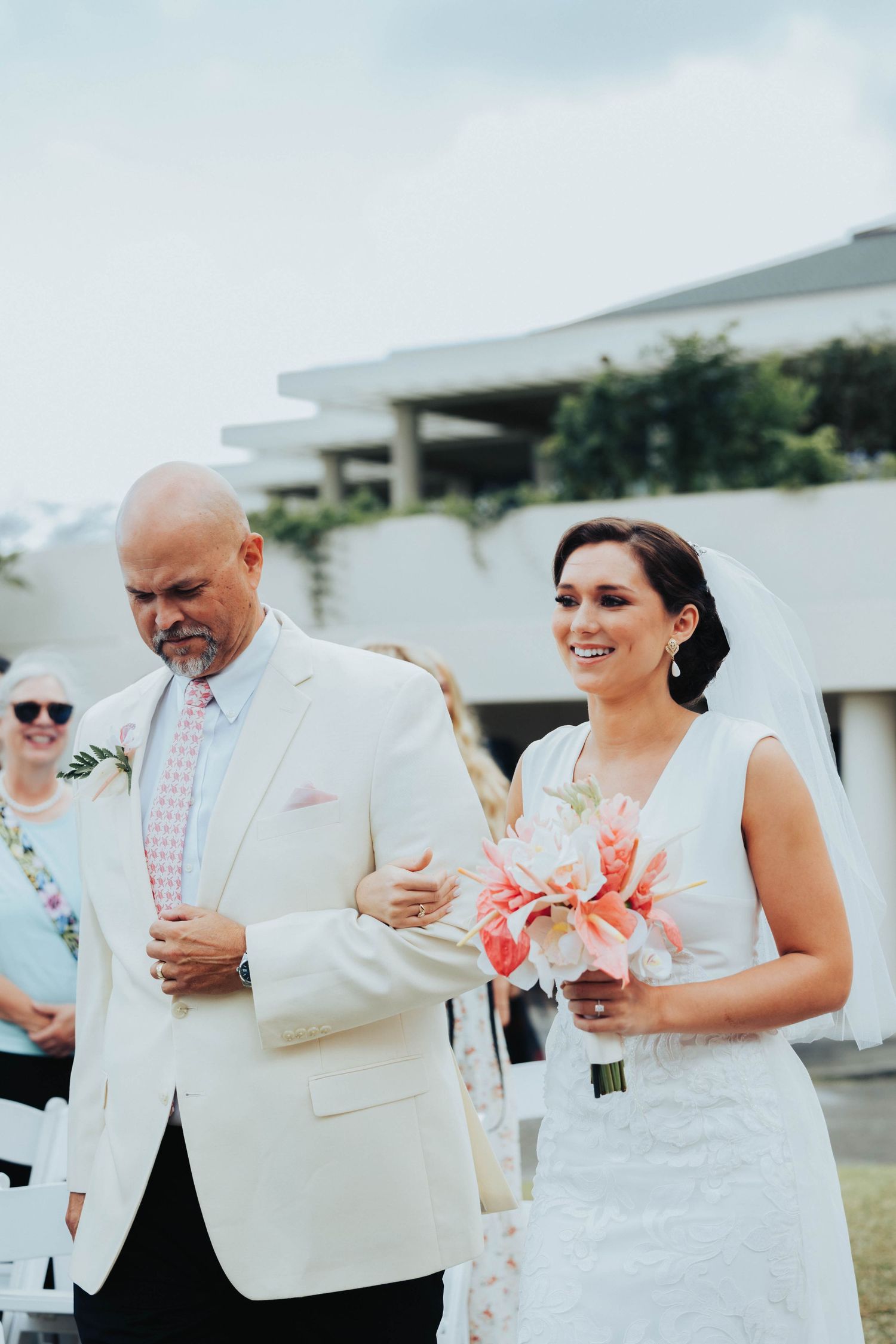 A couple walks arm in arm during an outdoor wedding ceremony, with the person in a white dress holding coral flowers.