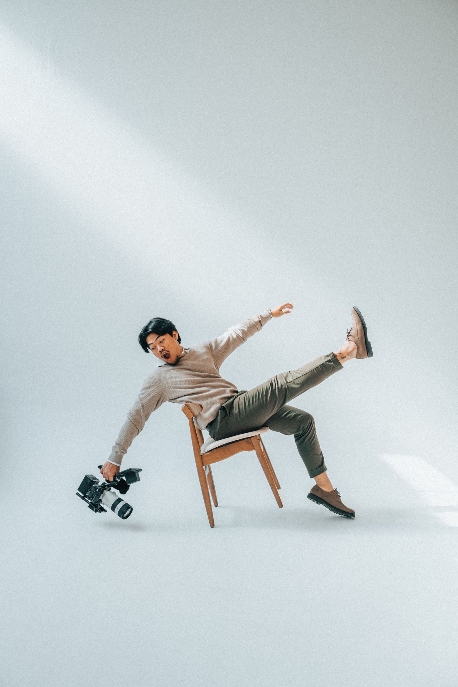 A dancer in casual attire performs a dynamic pose while balancing on a wooden chair against a white studio background.