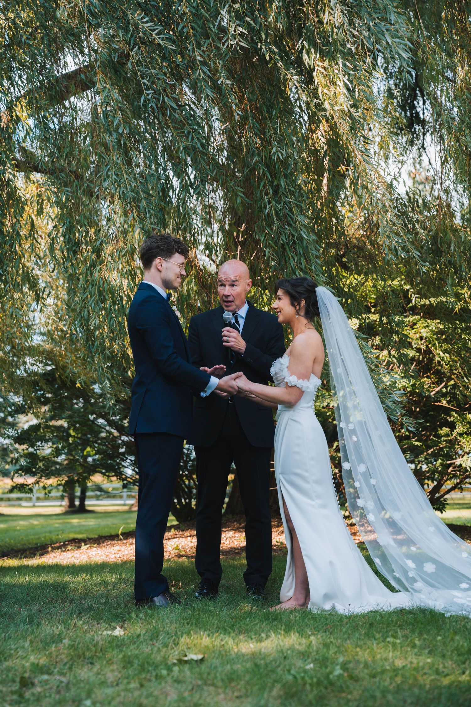 A romantic wedding ceremony takes place under a weeping willow tree in a lush outdoor garden setting.