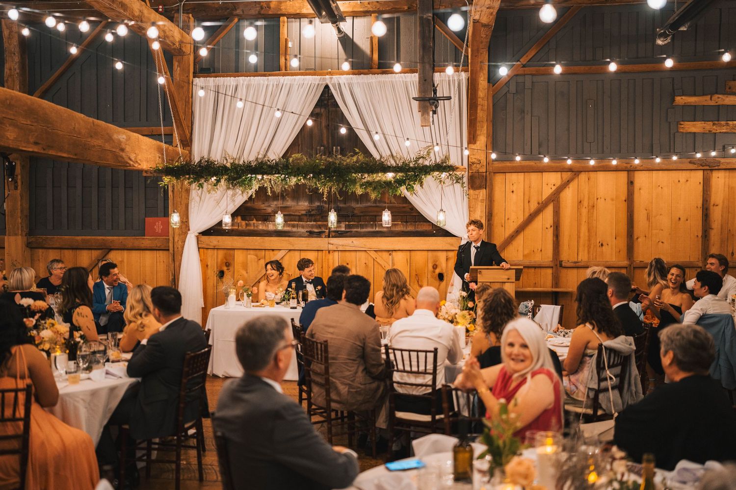 Wedding ceremony taking place inside a rustic barn venue with string lights and white drapes creating a romantic atmosphere.