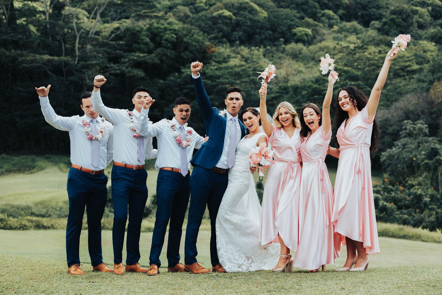 Wedding party in peach and navy attire poses playfully on a lush green lawn with trees in background.