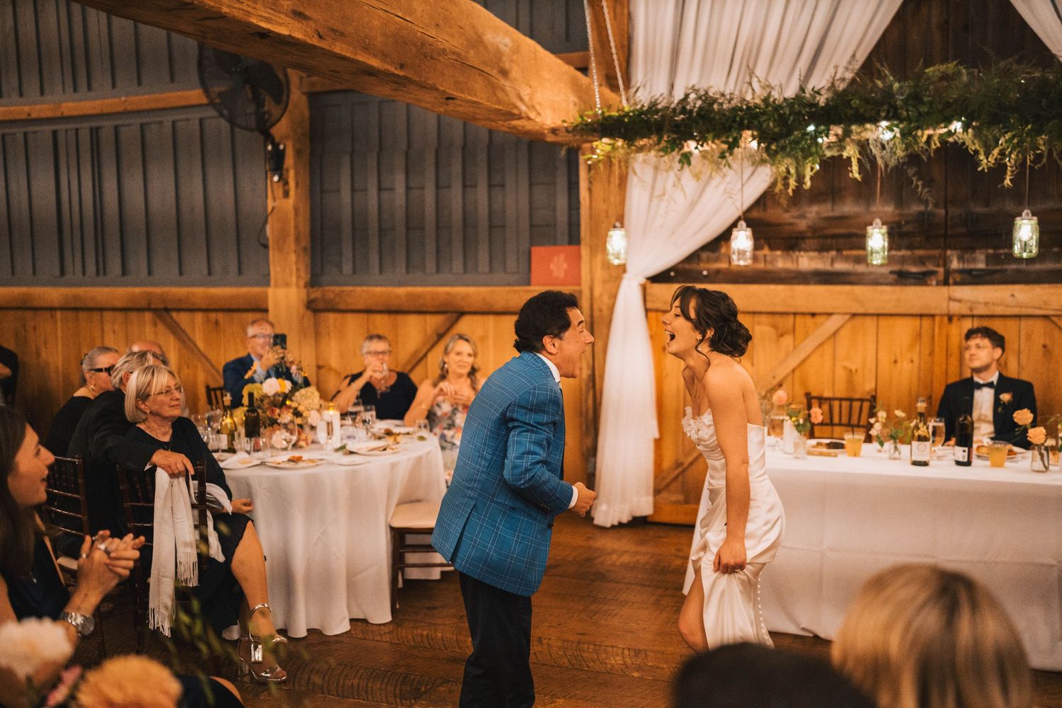 Romantic barn wedding reception with warm lighting, exposed wooden beams, and white fabric draping.