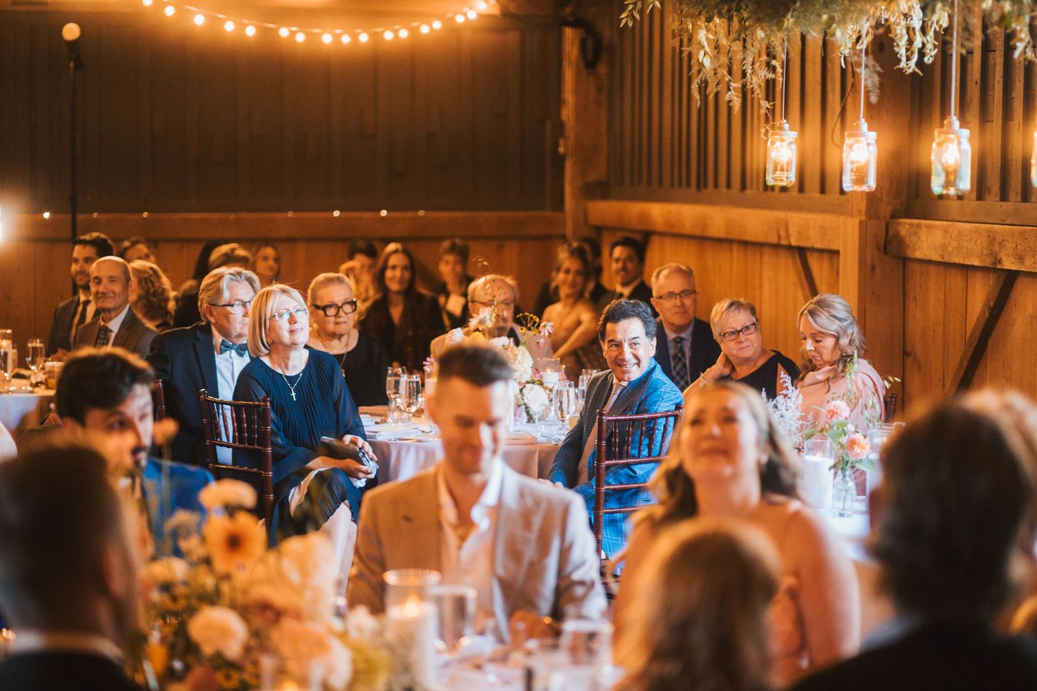 A rustic barn wedding reception with string lights and guests seated at long tables during dinner service.