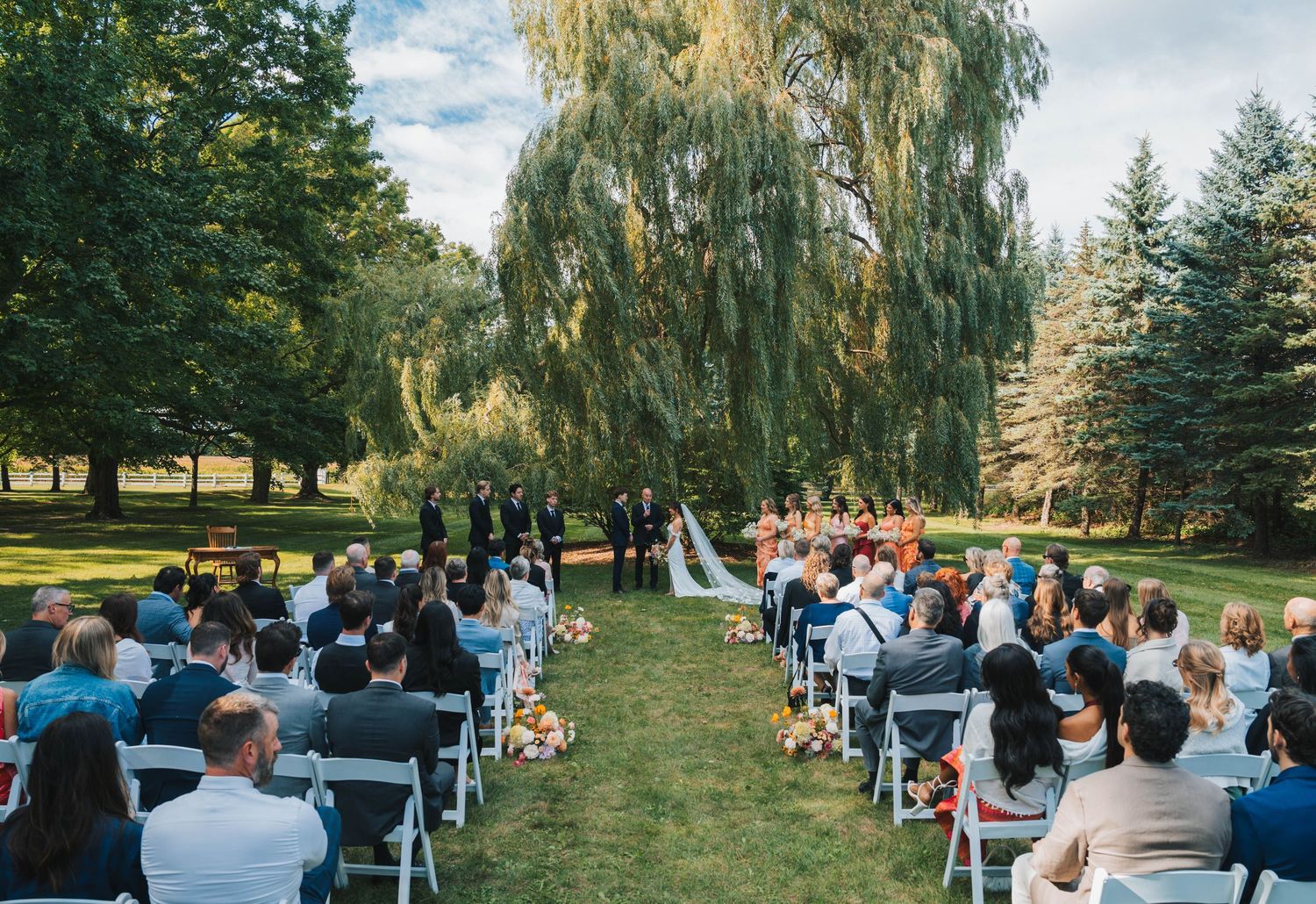 Wedding guests seated in rows on the lawn during an outdoor ceremony with tall trees in the background.