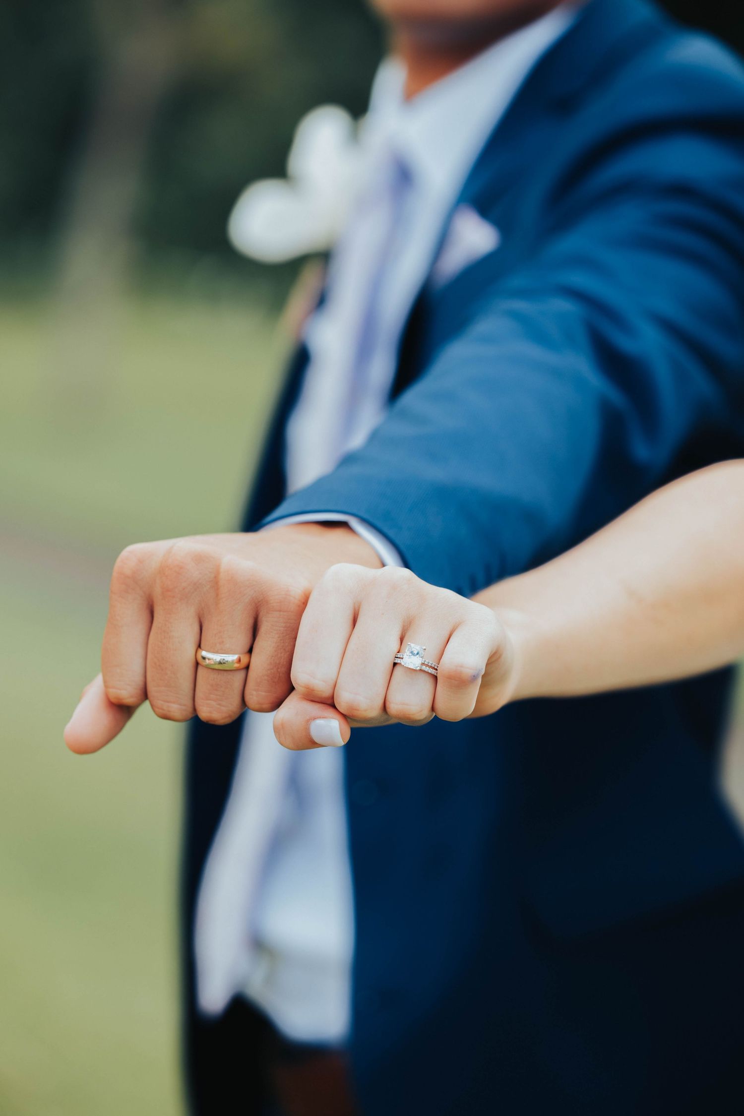 Close-up of newlyweds' hands showing wedding rings against blue suit background.