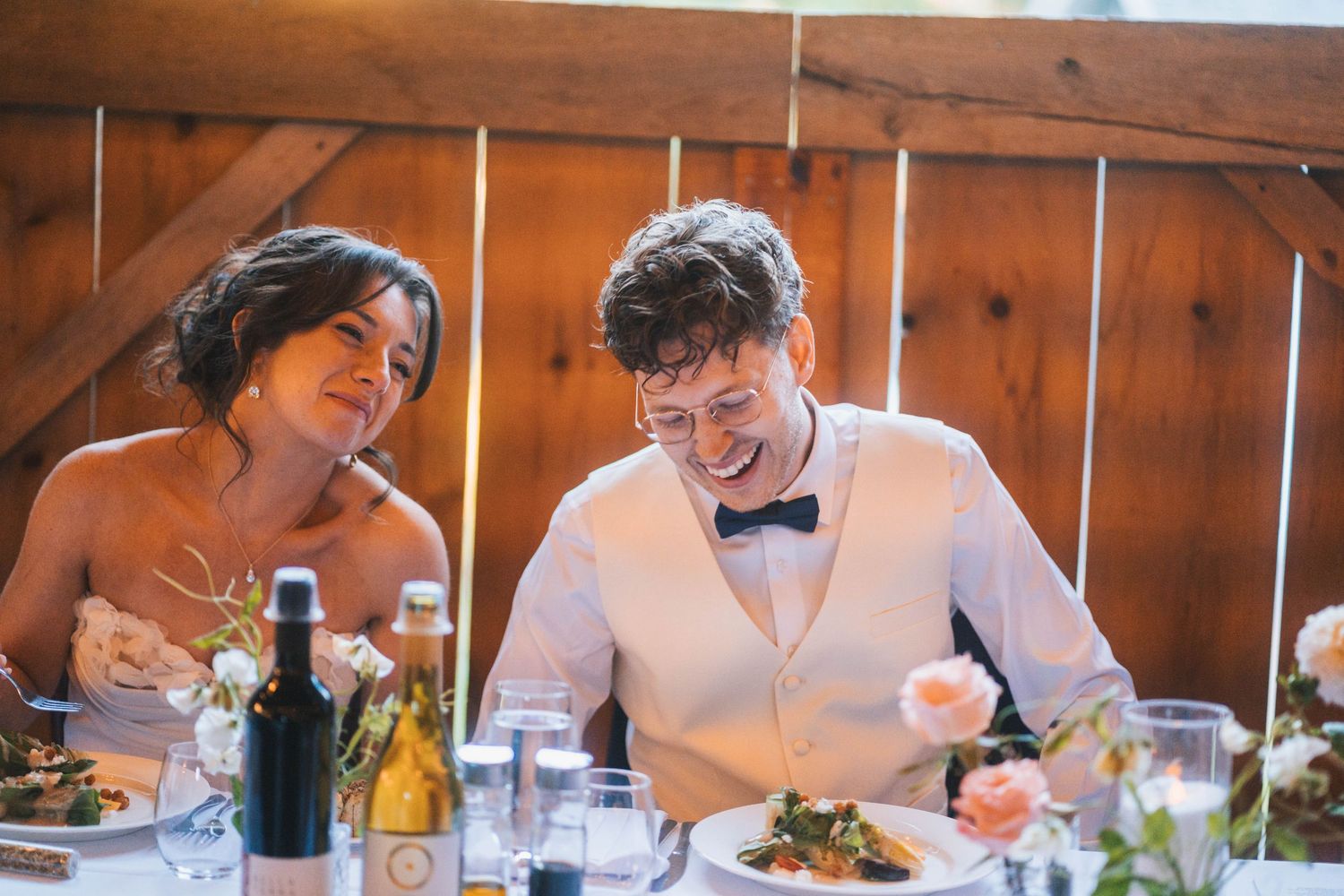 Wedding guests share joyful laughter while seated at a rustic wooden table during a reception celebration.