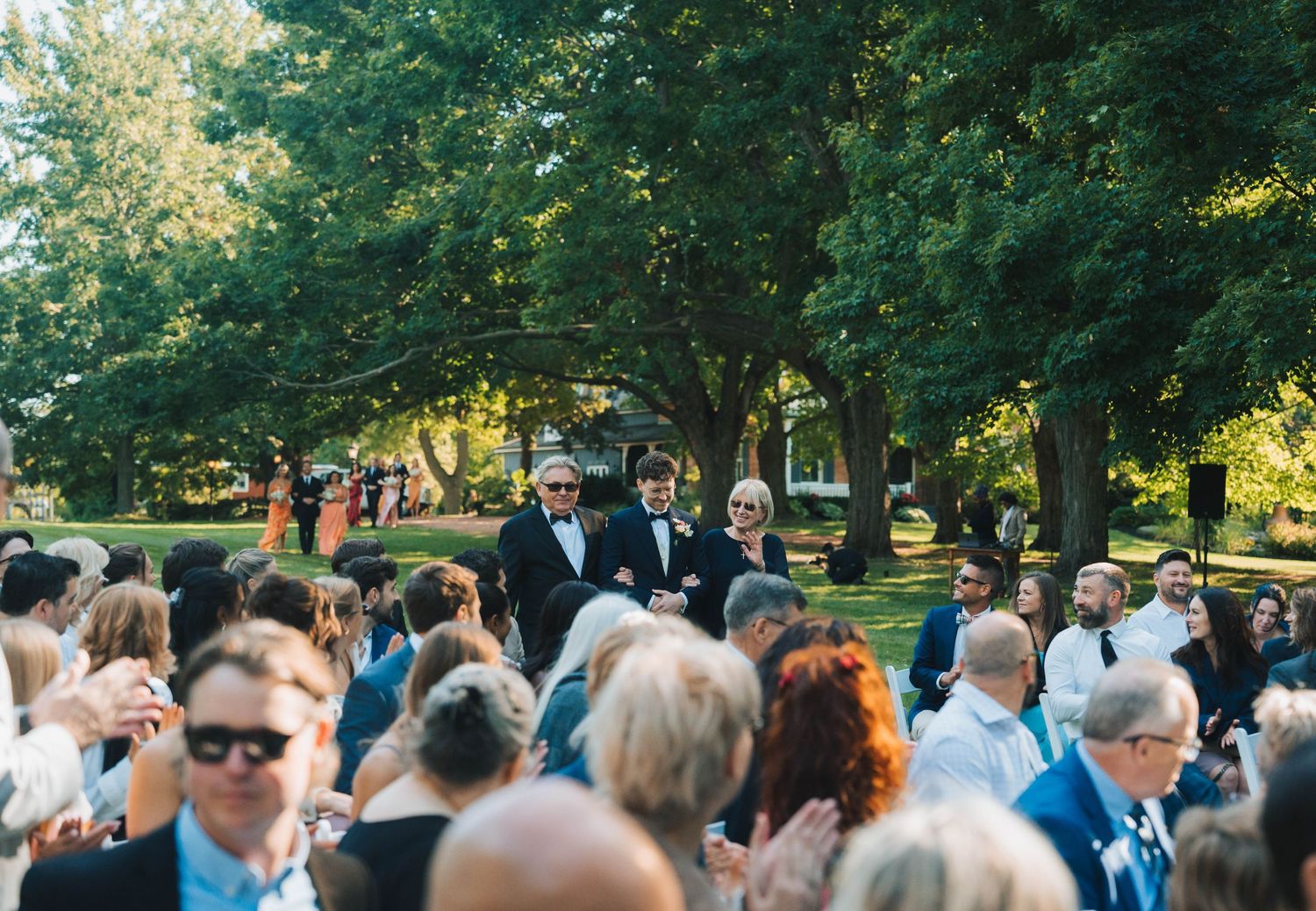 A large crowd gathers outdoors in a park-like setting during a sunny day event.