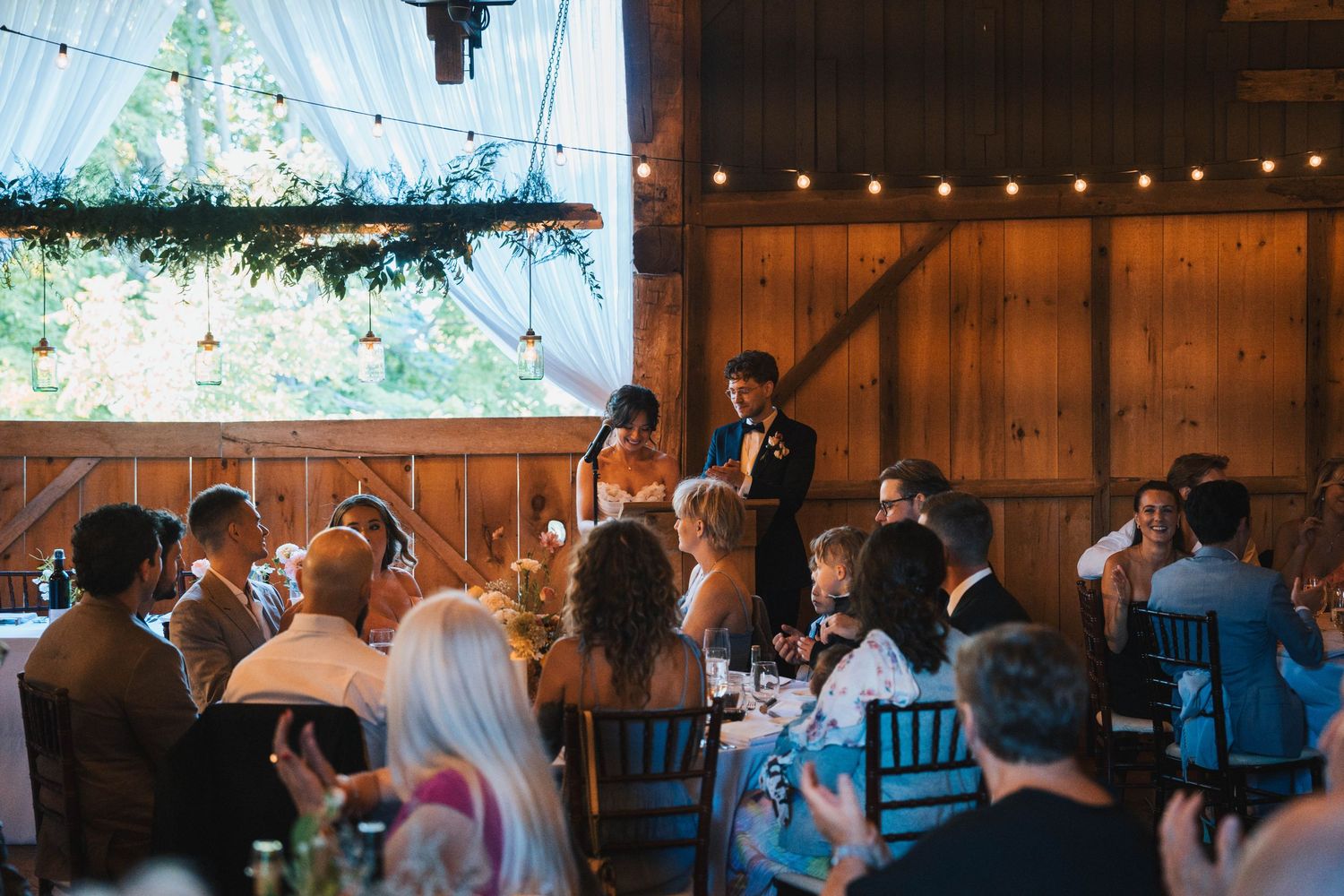 String lights illuminate a rustic barn wedding reception with guests seated at long wooden tables.