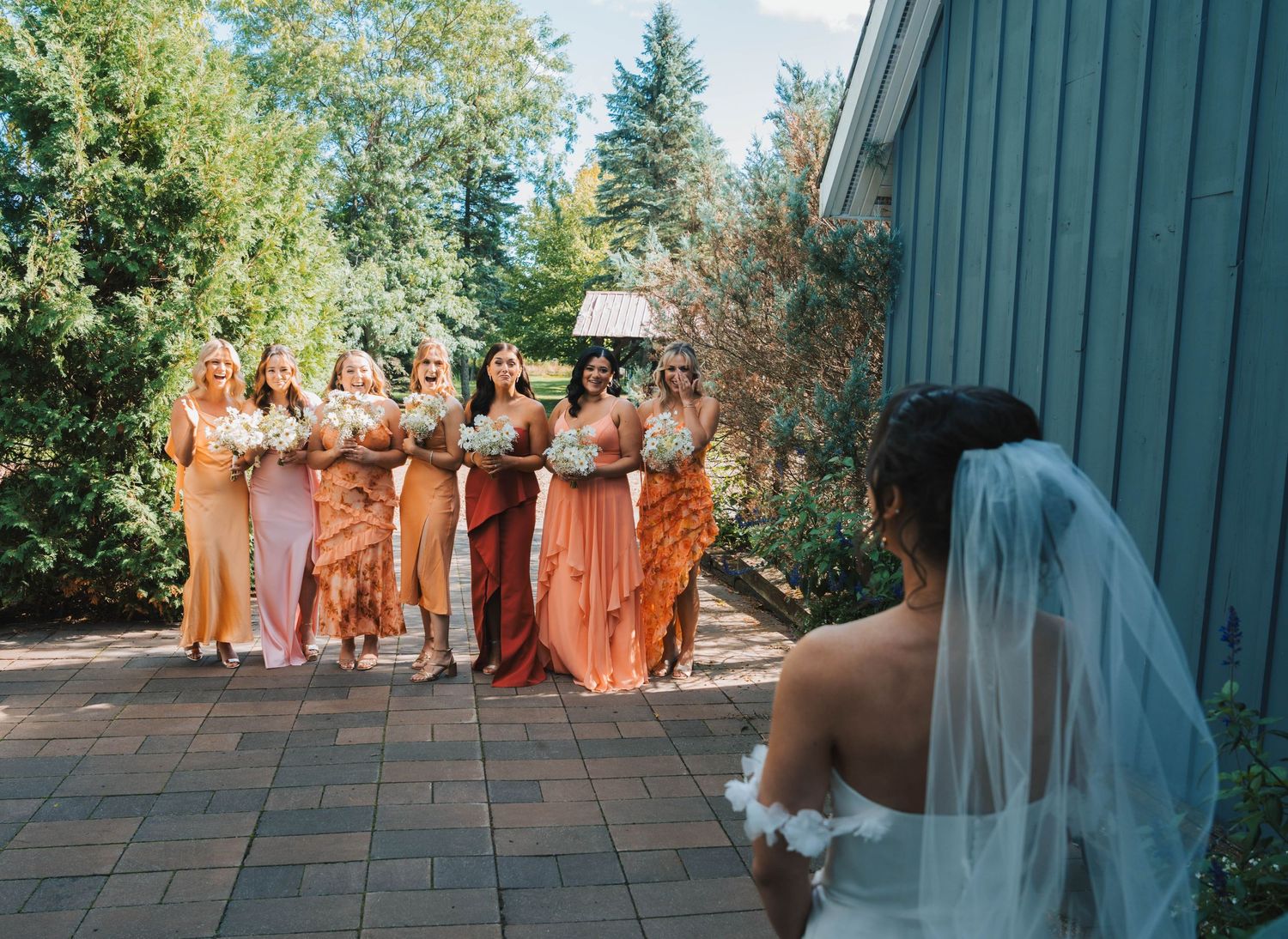 Wedding party in peach-colored dresses poses outdoors in front of evergreen trees and a blue building.