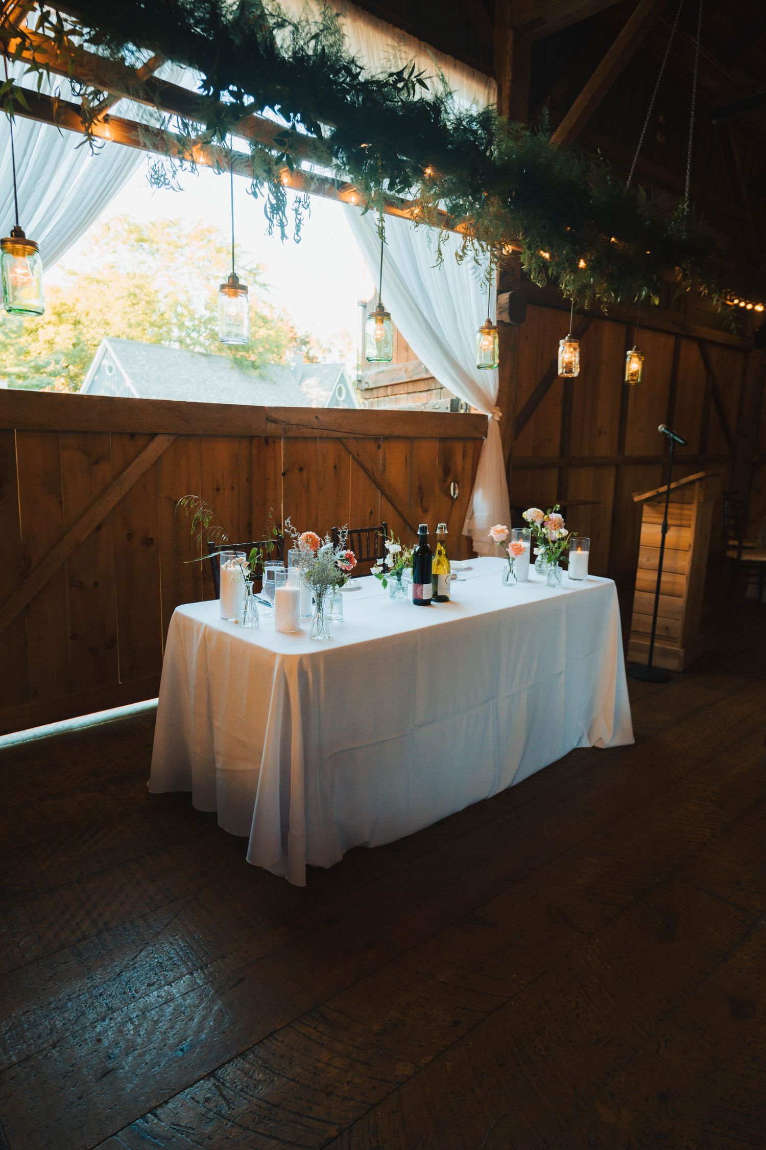 White-clothed tables with glassware and decorations line the rustic interior of a dimly lit wooden barn venue.