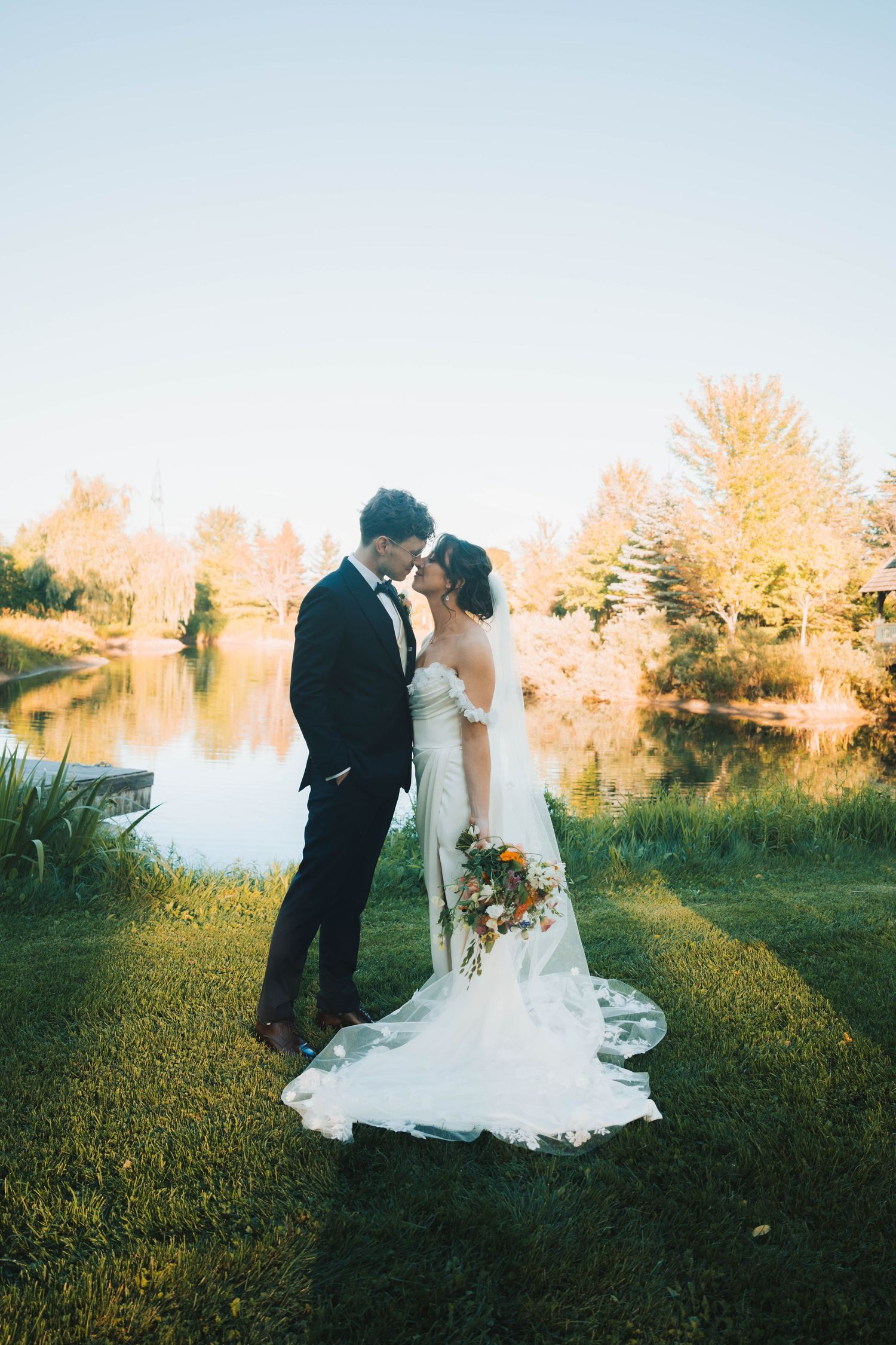 Newlywed couple shares romantic moments by a serene lake at sunset with autumn trees in the background.