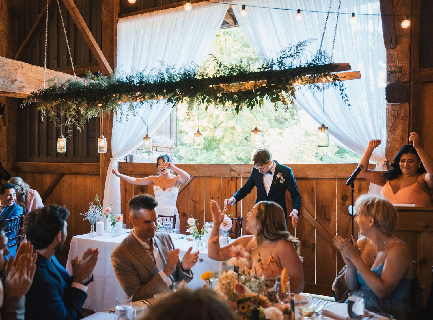 Wedding reception celebration inside a rustic wooden barn with string lights and greenery decorations overhead.