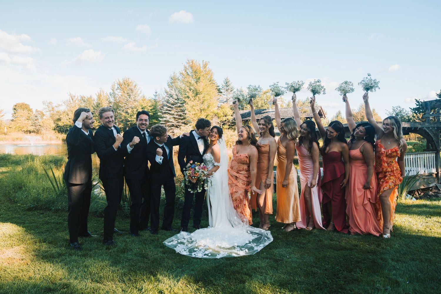 Wedding party stands on grass in autumn with bridesmaids in earth-toned dresses and groomsmen in black suits.