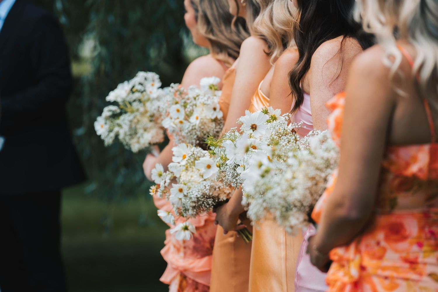 Bridesmaids in coral dresses hold white and cream floral bouquets during an outdoor wedding ceremony.