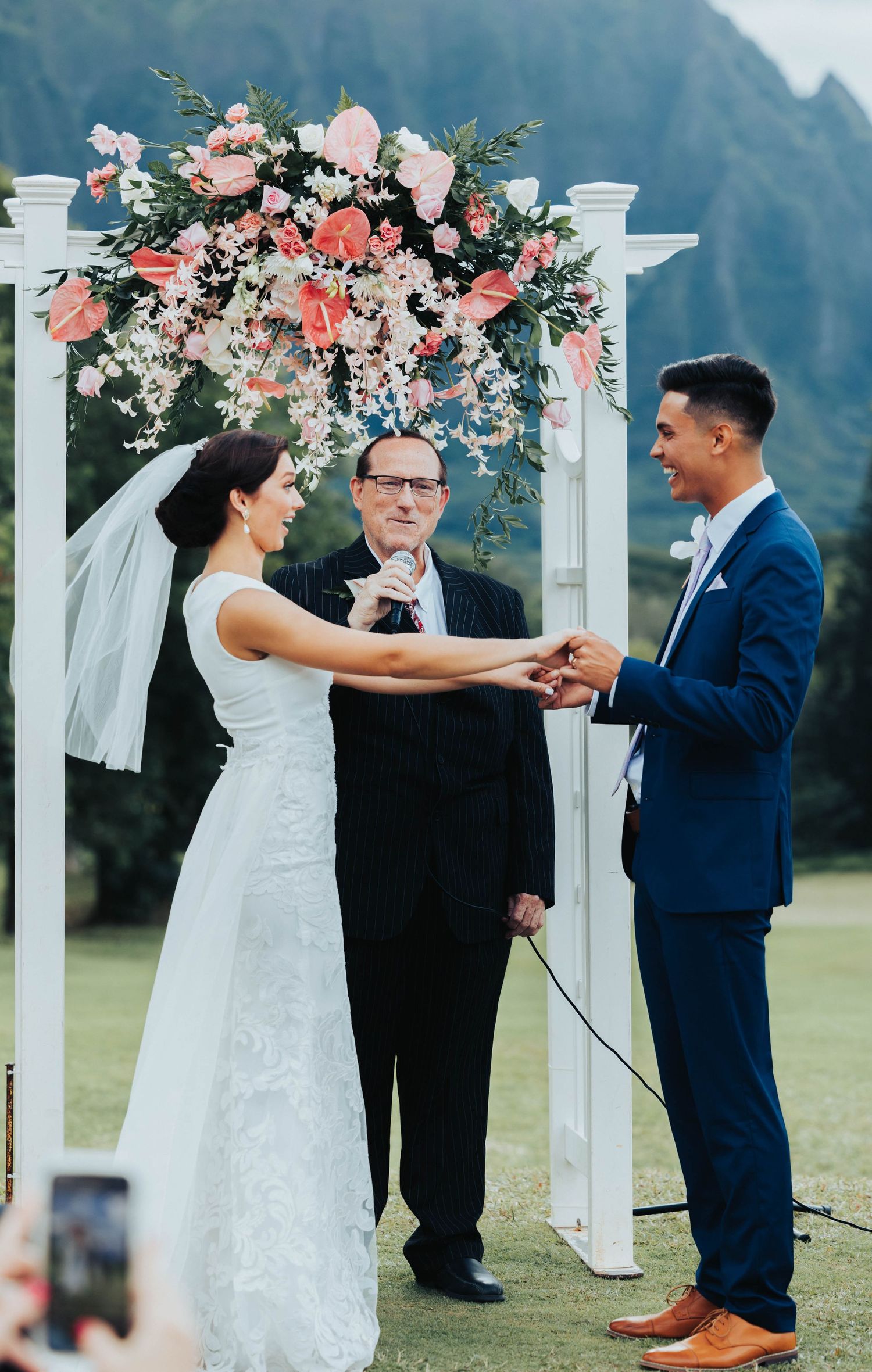 A wedding ceremony takes place under a white arch decorated with coral and pink flowers against a mountain backdrop.