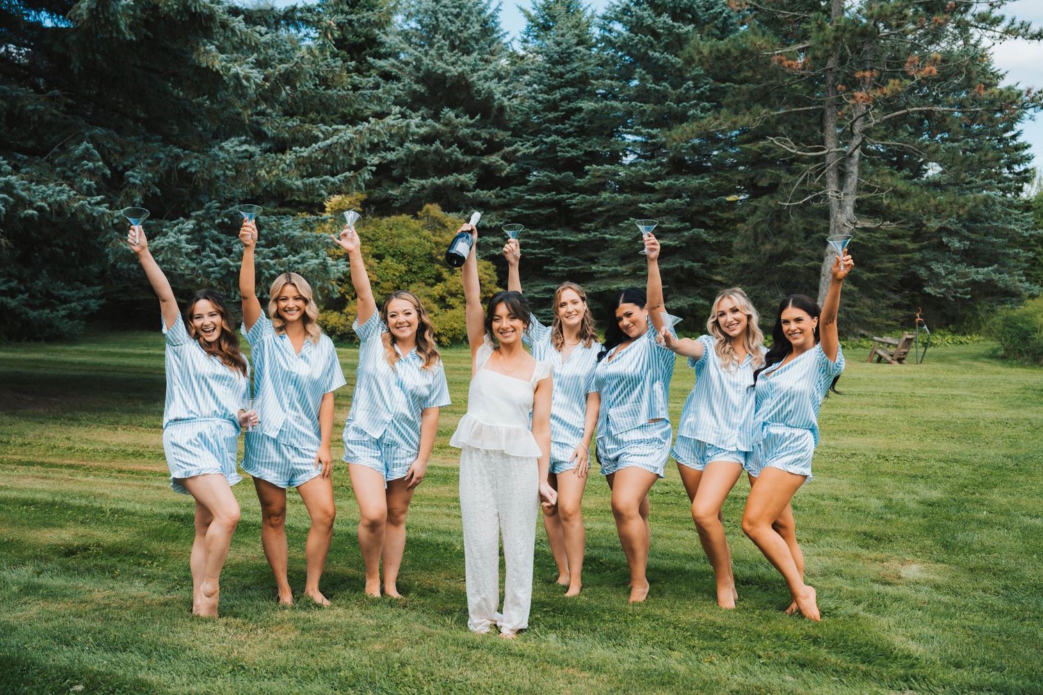 A group in matching light blue outfits poses joyfully on a lawn with evergreen trees in the background.